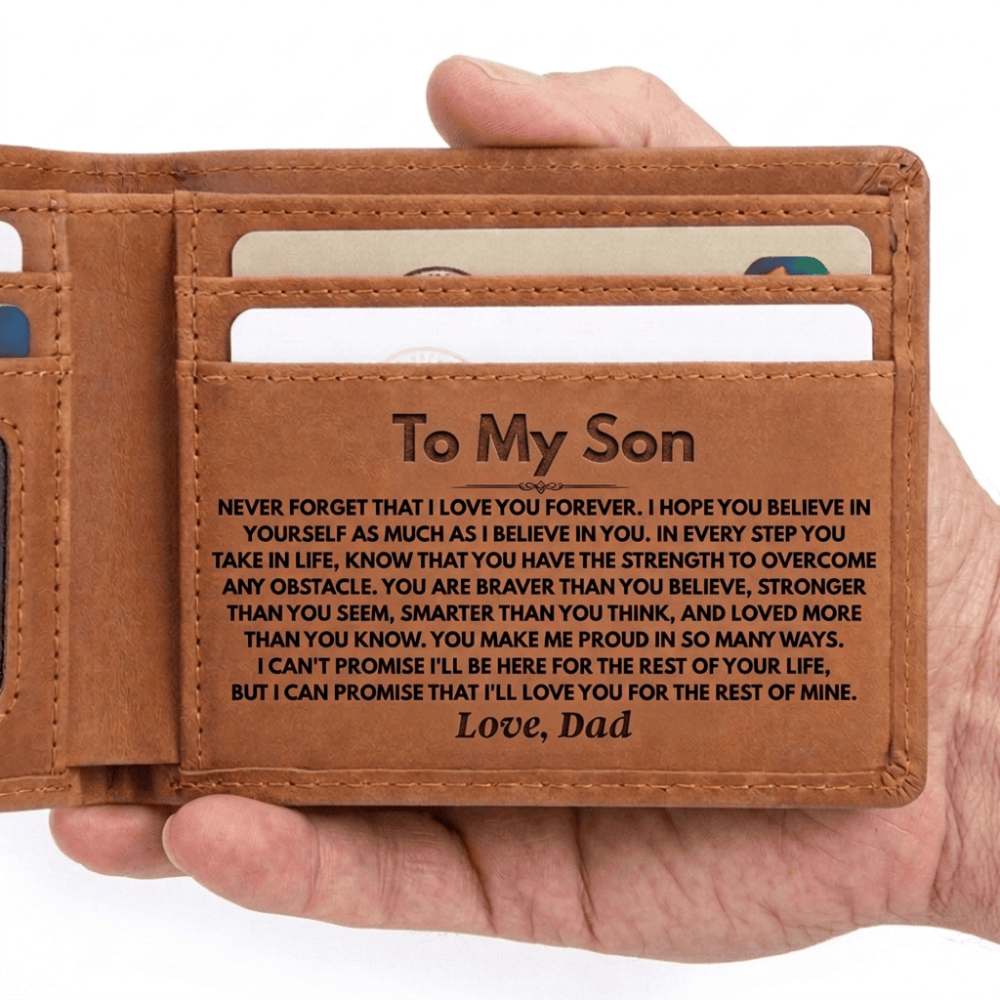 A close-up of a hand holding an open brown leather wallet, showing the interior card slots and a front pocket engraved with a sentimental message for a son.